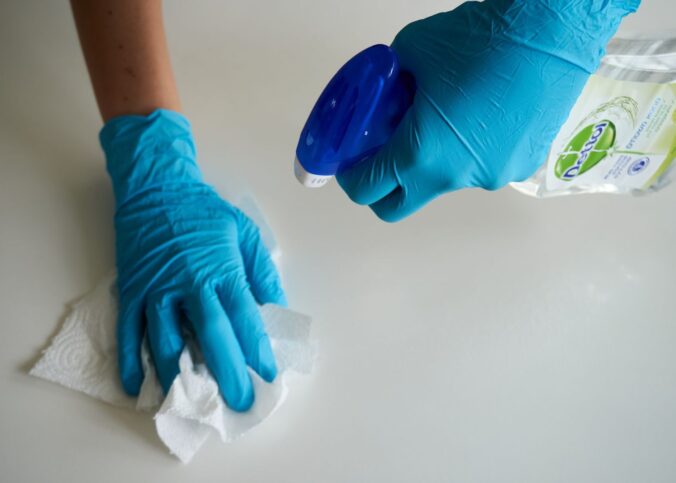 Close-up of hands wearing gloves cleaning a surface with disinfectant spray.