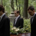 Three men in black suits carry a casket through a peaceful forest cemetery, symbolizing grief and togetherness.