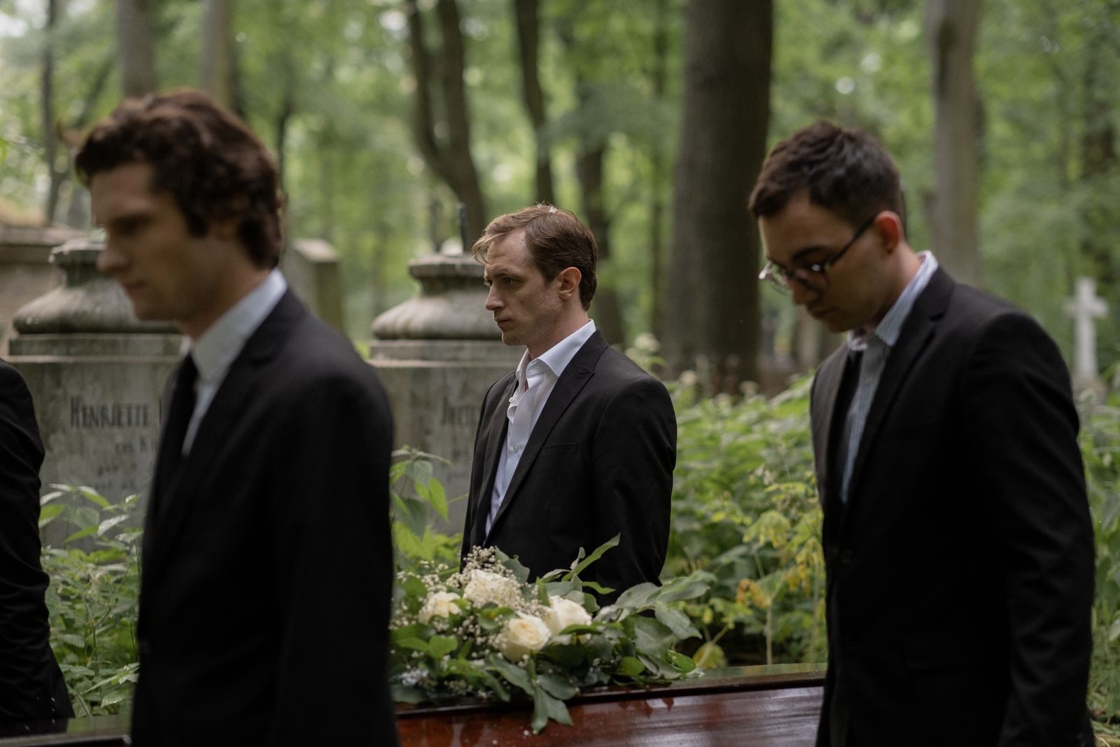 Three men in black suits carry a casket through a peaceful forest cemetery, symbolizing grief and togetherness.